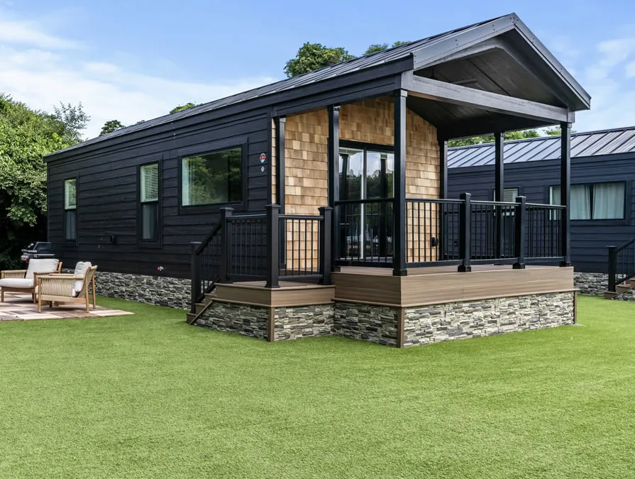 A park model cottage with a covered porch, cedar-shake accents and a stone skirt on a green lawn