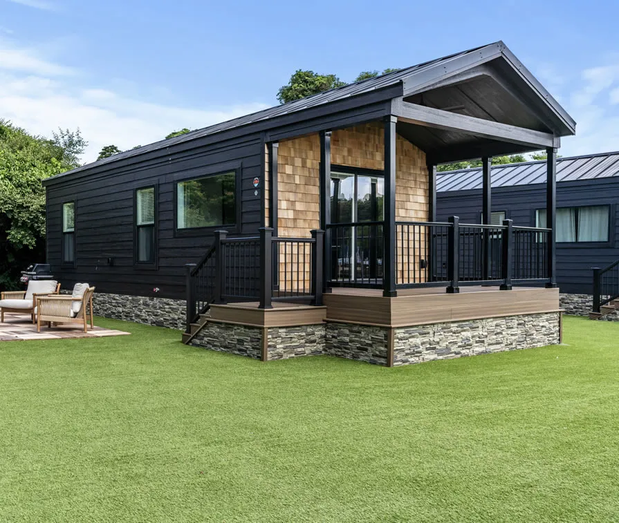 A park model cottage with a covered porch, cedar-shake accents and a stone skirt on a green lawn