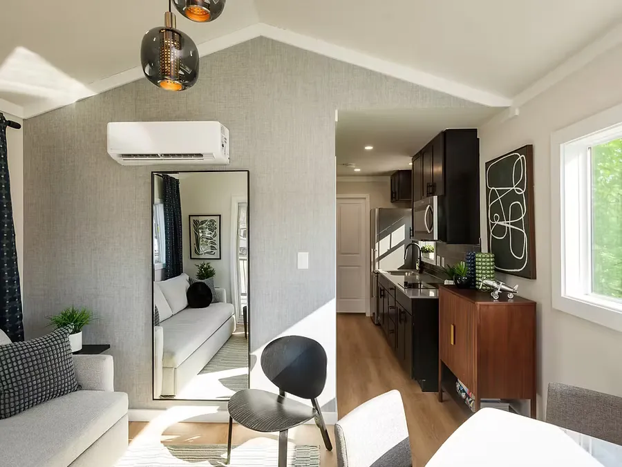 A bright cottage living room with a gray sofa, a tall mirror and a galley kitchen